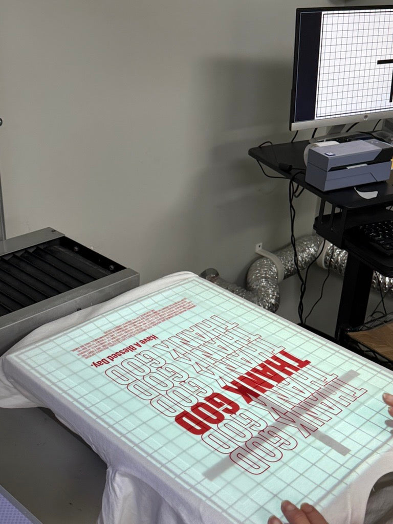 White T-shirt on a screen-printing press with a red “THANK GOD” graphic aligned under a projection grid during the production process.